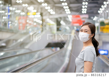 Beautiful Asian young woman with protective face mask looking and smiling to the camera while walking in the airport. Travel and tourist concept during the coronavirus or COVID-19 pandemic. Beautiful Asian young woman with protective face mask looking and smiling to the camera while walking in the airport. Travel and tourist concept during the coronavirus or COVID-19 pandemic. 86316217