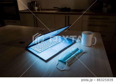 Remote work overtime during the quarantine period in the home office. Working atmosphere in the kitchen on a laptop in the dark late at night. Coffee mug and medical mask on the table. 86325839