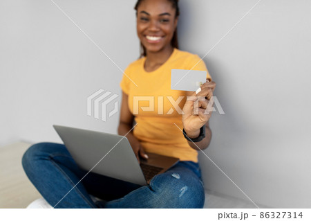Happy African American woman with laptop showing empty credit card at camera against grey studio wall, mockup 86327314