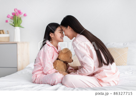 Happy asian teen girl with toy and millennial woman in pink pajamas enjoy time together sitting on bed 86327715