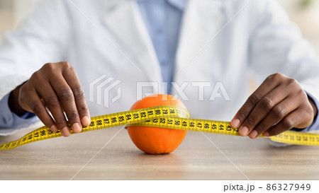 Cellulite is consequence of poor nutrition. African american nutritionist holding measuring tape around orange fruit 86327949