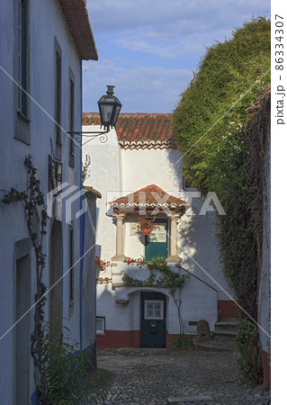 Narrow street of the ancient city of Obidos, Portugal. Scenic old town with medieval architecture. White houses is surrounded by greenery and red tiled roofs. 86334307