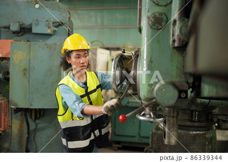 Female Industrial Worker in the Hard Hat with  Heavy Industry Manufacturing Factory in the Background with Various Metalwork Project Partss.Work at the Heavy Industry Manufacturing Factory. 86339344