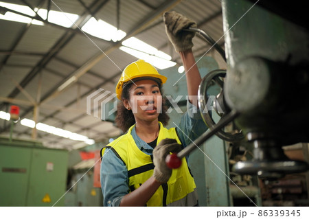 Female Industrial Worker in the Hard Hat with  Heavy Industry Manufacturing Factory in the Background with Various Metalwork Project Partss.Work at the Heavy Industry Manufacturing Factory. 86339345