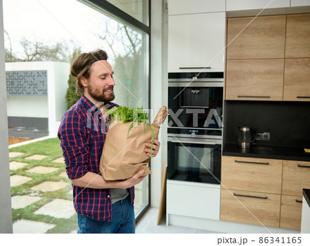 Charming young man enters the kitchen holding paper craft eco bag with food. Handsome guy with cardboard sack full of healthy food and whole grain bread, stands by kitchen countertop in spacious villa Charming young man enters the kitchen holding paper craft eco bag with food. Handsome guy with cardboard sack full of healthy food and whole grain bread, stands by kitchen countertop in spacious villa 86341165