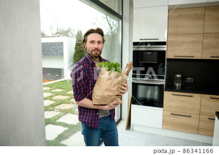 Handsome Caucasian man, charming guy, stands in a home kitchen with a cardboard bag full of healthy food and whole grain bread, looks at camera against the background of windows overlooking the garden Handsome Caucasian man, charming guy, stands in a home kitchen with a cardboard bag full of healthy food and whole grain bread, looks at camera against the background of windows overlooking the garden 86341166