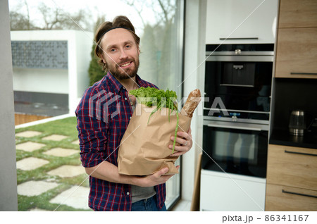 Happy charming Caucasian young man standing in home kitchen with eco paper bag full of healthy food and whole grain bread looking at camera against windows overlooking garden. Healthy eating concept Happy charming Caucasian young man standing in home kitchen with eco paper bag full of healthy food and whole grain bread looking at camera against windows overlooking garden. Healthy eating concept 86341167