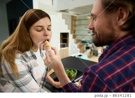 Happy heterosexual Caucasian couple in love enjoying cooking a healthy meal together in the kitchen island at home. Loving husband feeding his pregnant wife with a slice of yellow bell pepper Happy heterosexual Caucasian couple in love enjoying cooking a healthy meal together in the kitchen island at home. Loving husband feeding his pregnant wife with a slice of yellow bell pepper 86341281