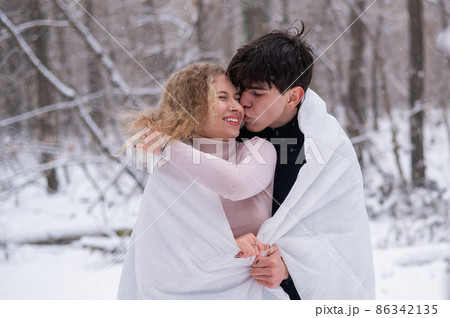 A young couple walks in the park in winter. The guy and the girl are kissing wrapped in a white blanket outdoors. A young couple walks in the park in winter. The guy and the girl are kissing wrapped in a white blanket outdoors. 86342135