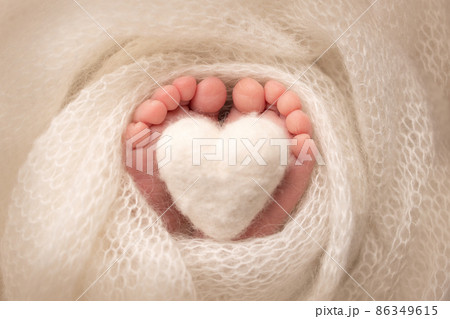 The tiny foot of a newborn baby. Soft feet of a new born in a white wool blanket. Close up of toes, heels and feet of a newborn. Knitted white heart in the legs of a baby. Macro photography.  86349615