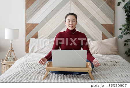 Relaxed mixed race young asian woman sitting on her bed with folded in mudra gesture hands, reducing stress during workday, chilling or doing yoga breathing exercises, working from home with laptop Relaxed mixed race young asian woman sitting on her bed with folded in mudra gesture hands, reducing stress during workday, chilling or doing yoga breathing exercises, working from home with laptop 86350907