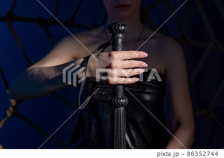 BDSM concept. A woman in a leather dress holds a black whip with a long fringe. Role-playing games for adults. Close-up of sex toys for domination. 86352744