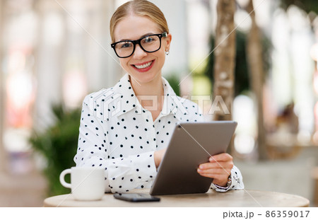 Lovely student girl in glasses sitting with tablet and cup of takeaway coffee outside on terrace Lovely student girl in glasses sitting with tablet and cup of takeaway coffee outside on terrace 86359017