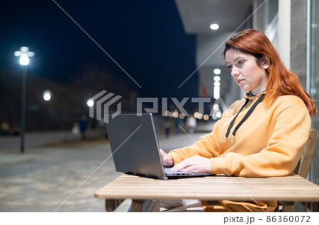 A woman works remotely at a laptop in a summer cafe late in the evening. Serious Girl studying while sitting on an empty street at a wooden table. Female freelancer in a sweatshirt. A woman works remotely at a laptop in a summer cafe late in the evening. Serious Girl studying while sitting on an empty street at a wooden table. Female freelancer in a sweatshirt. 86360072