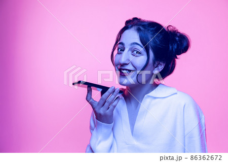 Studio shot of young beautiful girl in white blouse using smartphone isolated on pink background in neon light. Concept of emotions 86362672