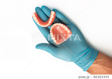 The doctor's hand in a blue glove holds a denture. Dental prosthesis in the hands of the doctor close-up. Dentist holding ceramic dental bridge. Dentistry conceptual photo. Prosthetic dentistry. 86363445