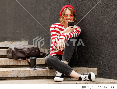 Smiling student girl using mobile phone while sitting outside with takeaway coffee before in morning 86367133