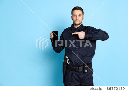 Portrait of young man, policeman officer wearing black uniform using phone isolated on blue background. Concept of job, caree, law and order. Portrait of young man, policeman officer wearing black uniform using phone isolated on blue background. Concept of job, caree, law and order. 86370150