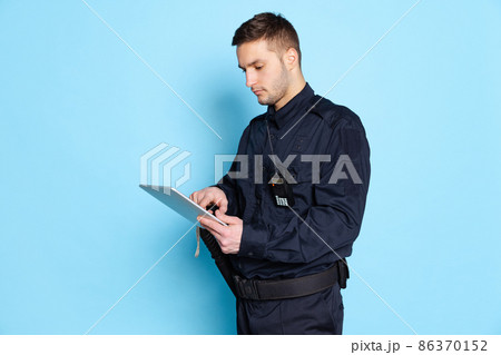 Portrait of young man, policeman officer wearing black uniform using tablet isolated on blue background. Concept of job, caree, law and order. Portrait of young man, policeman officer wearing black uniform using tablet isolated on blue background. Concept of job, caree, law and order. 86370152