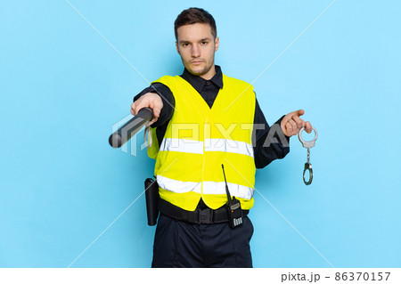 Half-length portrait of young male policeman officer wearing black uniform and warnvest posing isolated on blue background. Concept of job, caree, law and order. Half-length portrait of young male policeman officer wearing black uniform and warnvest posing isolated on blue background. Concept of job, caree, law and order. 86370157