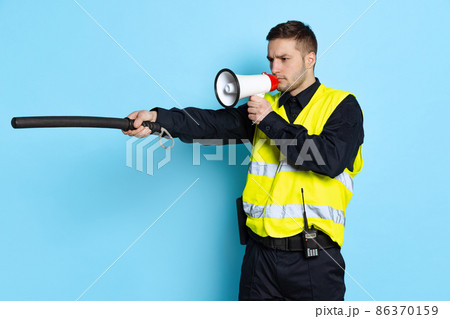 Portrait of young policeman officer wearing black uniform and warnvest shouting at megaphone isolated on blue background. Concept of job, caree, law and order. Portrait of young policeman officer wearing black uniform and warnvest shouting at megaphone isolated on blue background. Concept of job, caree, law and order. 86370159