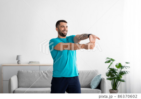 Smiling happy millennial european guy in blue t-shirt doing exercises and stretching for arms 86370602