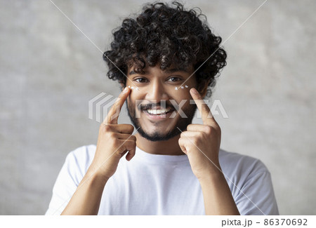 Portrait of happy indian man applying cream under eyes and smiling at camera, enjoying morning beauty routine Portrait of happy indian man applying cream under eyes and smiling at camera, enjoying morning beauty routine 86370692