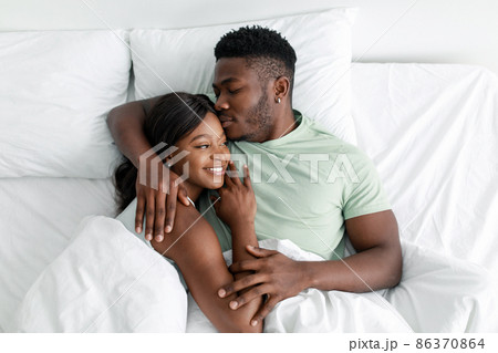 Cheerful young african american husband hugging wife and kissing on comfortable bed, resting, relax in bedroom 86370864