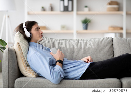 Rest and relax concept. Calm guy lying on sofa, listening to music, enjoying meditation for sleep in wireless headphones Rest and relax concept. Calm guy lying on sofa, listening to music, enjoying meditation for sleep in wireless headphones 86370873