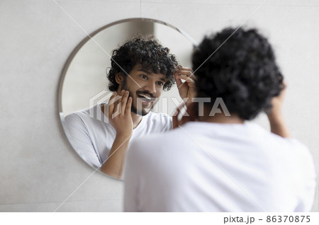 Male beauty and haircare routine. Handsome indian man touching beard and hair, looking in mirror in bathroom 86370875