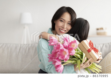 Little Asian girl hugging granny, presenting her with flowers and gift for Women's Day, congratulating with birthday Little Asian girl hugging granny, presenting her with flowers and gift for Women's Day, congratulating with birthday 86370995