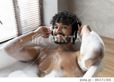 Calm indian man in wireless headphones lying in hot bubbly bath and enjoying his playlist in bathroom at home 86371366