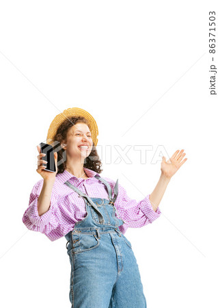 Excited young girl, female gardener in work uniform and hat using phone isolated on white background. Concept of job, emotions, agronomy, eco 86371503