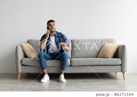 Cheerful Arab man talking on cellphone sitting on couch 86371504