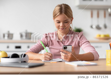 Smiling caucasian adolescent girl pupil makes notes and watch at smartphone, check lesson at table in kitchen 86371526