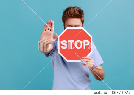 Portrait of bearded man covering half of face with octagonal Stop symbol, showing red traffic sign and ban gesture with palm, warning of forbidden way. Indoor studio shot isolated on blue background. 86373027