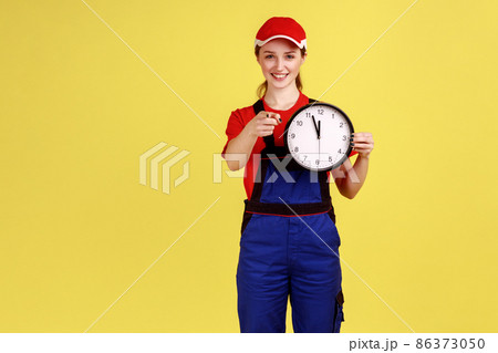 Portrait of worker woman standing with big wall clock in hands, looking and pointing at camera with smile, wearing overalls and red cap. Indoor studio shot isolated on yellow background. 86373050