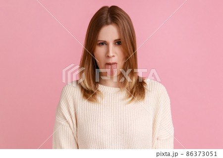 Portrait of blond woman sticking out tongue and looking at camera, teasing with naughty expression, disobedient behavior, wearing white sweater. Indoor studio shot isolated on pink background. 86373051