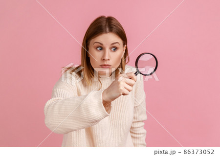 Portrait of blond woman looking through magnifying glass, spying, finding out something, exploring crime scene, inspecting, wearing white sweater. Indoor studio shot isolated on pink background. 86373052