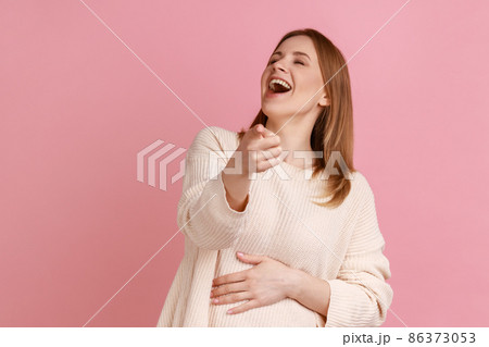 Portrait of optimistic blond woman laughing out loud pointing finger, making fun of you and looking at camera, joke, wearing white sweater. Indoor studio shot isolated on pink background. 86373053