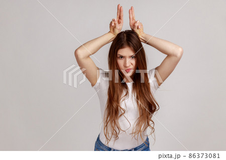 Portrait of aggressive bully woman with dark hair showing bull horns gesture over head, frowning as before attack, wearing white T-shirt. Indoor studio shot isolated on gray background. 86373081