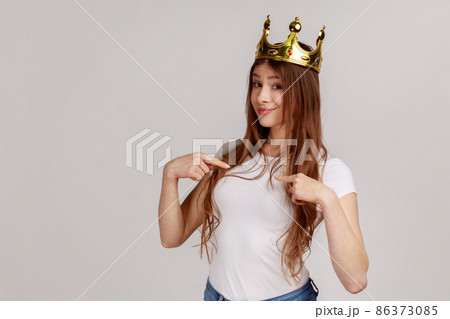 Portrait of selfish egoistic woman with golden crown on head pointing herself and looking at camera, declaring her authority, wearing white T-shirt. Indoor studio shot isolated on gray background. 86373085
