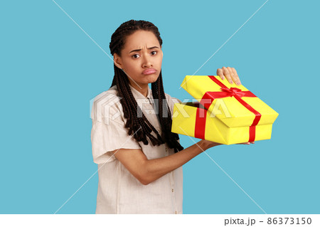 Portrait of unhappy upset woman with black dreadlocks looking into gift box, opening present and looking at camera with sorrow, wearing white shirt. Indoor studio shot isolated on blue background. Portrait of unhappy upset woman with black dreadlocks looking into gift box, opening present and looking at camera with sorrow, wearing white shirt. Indoor studio shot isolated on blue background. 86373150