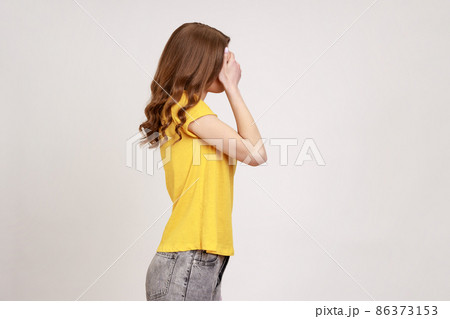I'm not watching. Profile portrait of slim girl in yellow casual T- shirt covering eyes with hand, frightened to look, sees disgusting things. Indoor studio shot isolated on gray background. 86373153