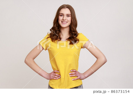 Young female with brown wavy hair posing with hands on hips and looking directly at camera with positive and happy expression, wears yellow T-shirt. Indoor studio shot isolated on gray background. 86373154