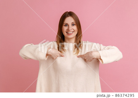 Portrait of blond woman pointing herself and looking at camera with toothy smile, proud of own success, achievement, wearing white sweater. Indoor studio shot isolated on pink background. 86374295