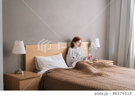 Remote view of happy young woman enjoy shopping online with credit card and mobile phone lying in bed at home. Smiling female holding credit card to make online payment at bedroom. Remote view of happy young woman enjoy shopping online with credit card and mobile phone lying in bed at home. Smiling female holding credit card to make online payment at bedroom. 86374319