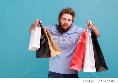 Portrait of tiered exhausted handsome bearded man raised arms with shopping bags,looking at camera, feels bored after going in mall. Indoor studio shot isolated on blue background. Portrait of tiered exhausted handsome bearded man raised arms with shopping bags,looking at camera, feels bored after going in mall. Indoor studio shot isolated on blue background. 86374407