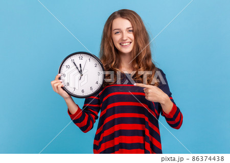 Winsome woman wearing striped casual style sweater, pointing big clock and looking at camera with positive, showing wall watches to hurry up. Indoor studio shot isolated on blue background. Winsome woman wearing striped casual style sweater, pointing big clock and looking at camera with positive, showing wall watches to hurry up. Indoor studio shot isolated on blue background. 86374438