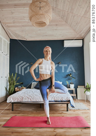 Smiling woman in sportswear does vrksasana holding leg in spacious room Smiling woman in sportswear does vrksasana holding leg in spacious room 86376700